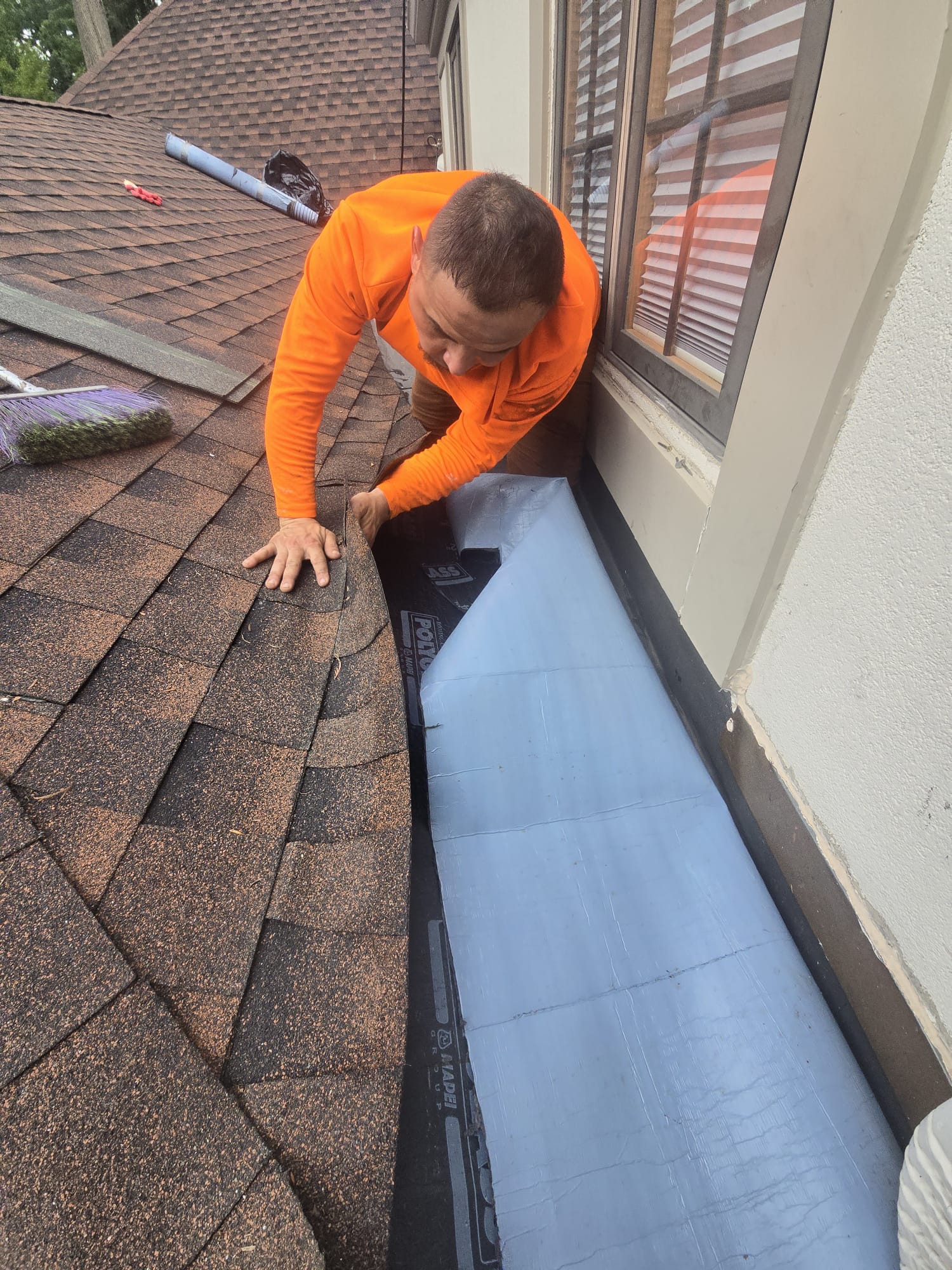 Roofing worker in an orange shirt repairing shingles next to a house window